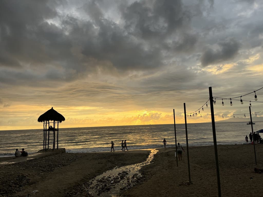 A beach volleyball net and a thatched lifeguard hut at sunset, a few people on the sand