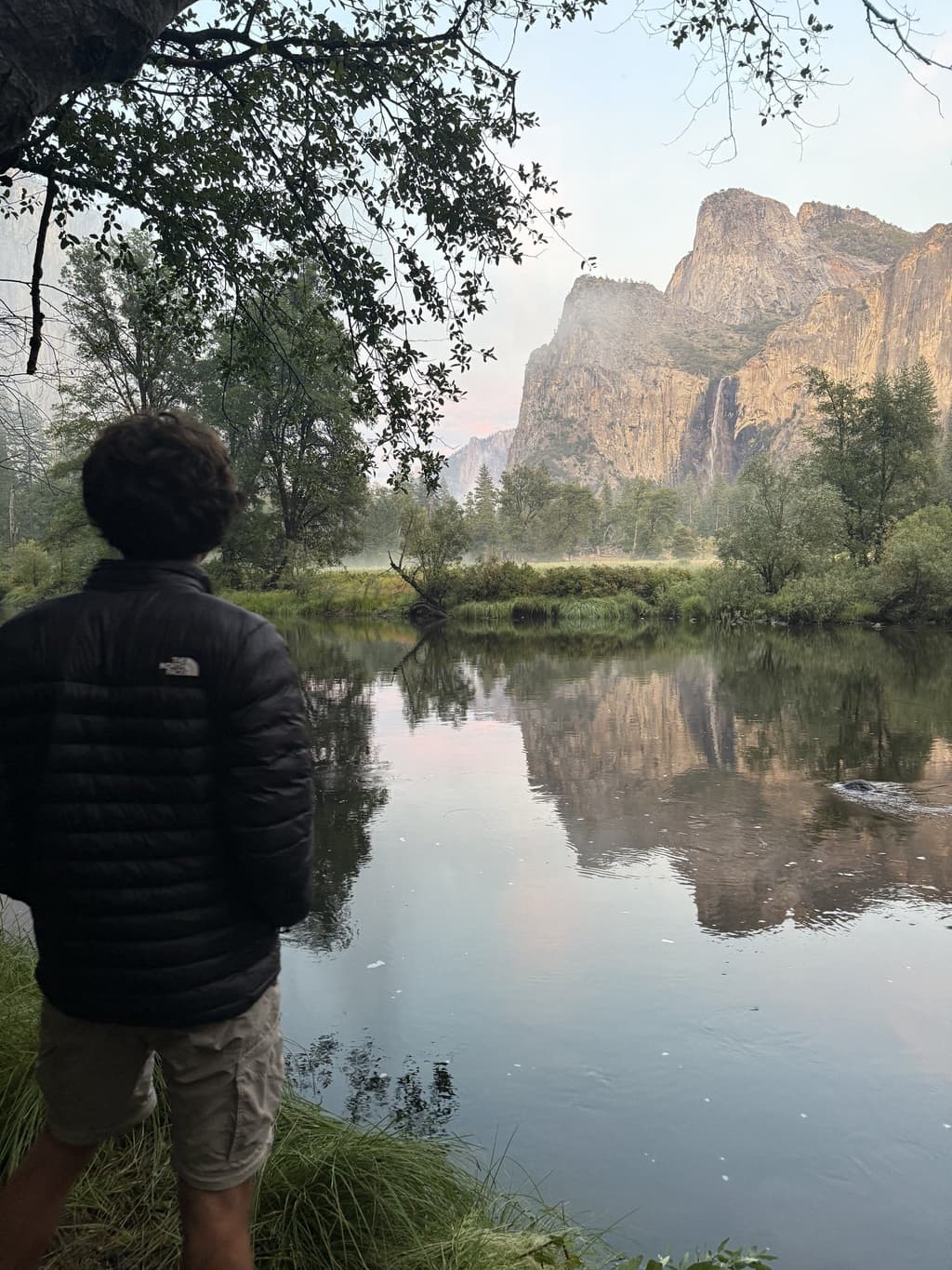 Kyle from behind in a puffer jacket, looking across a still river at Yosemite's cliffs